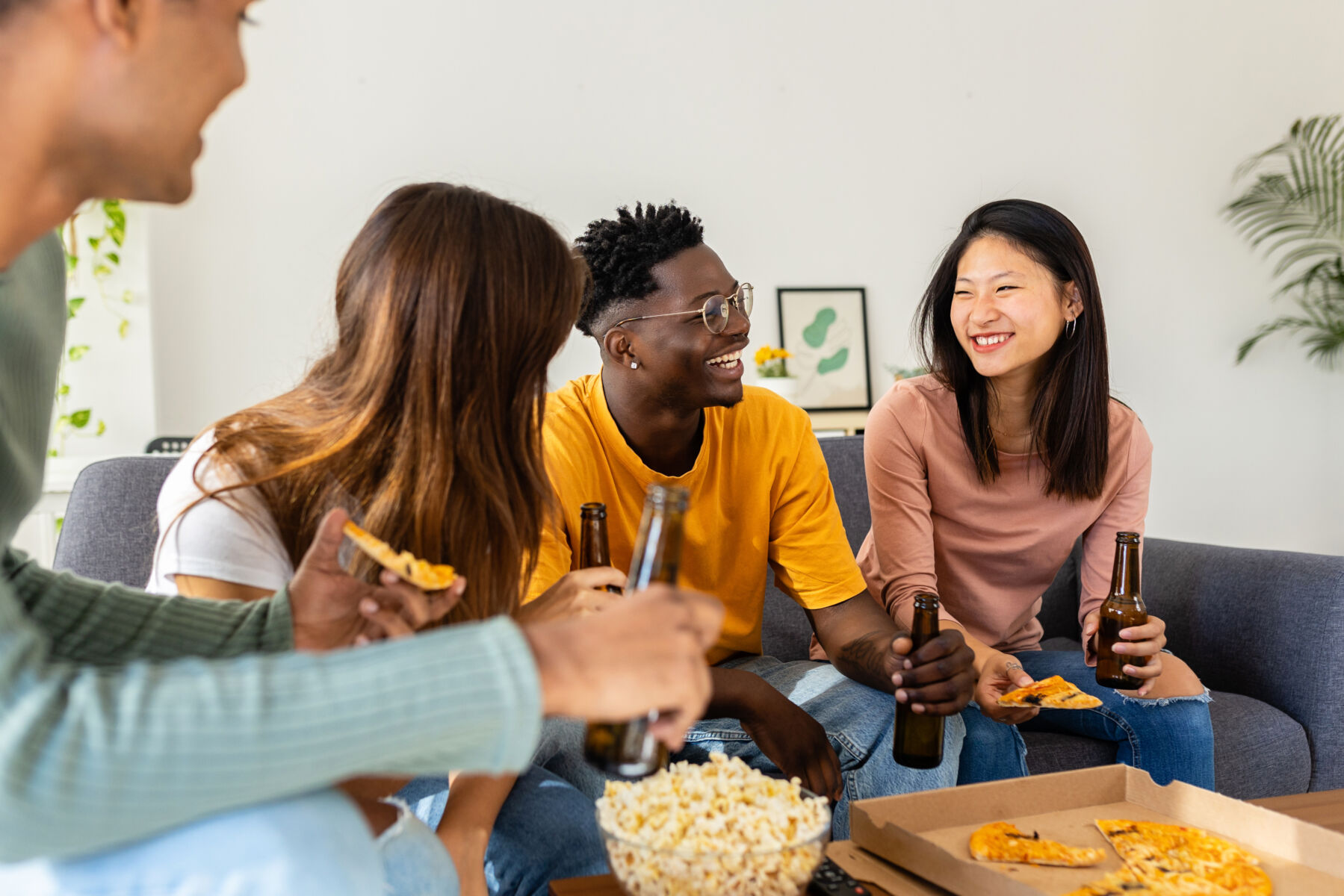 Group of friends having fun at lunch eating pizza and drinking beer. Young diverse people spending time together laughing sitting on sofa at home. International friendship concept