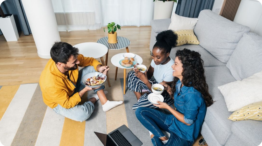 A man and two women sit on the floor of a living room while eating food.